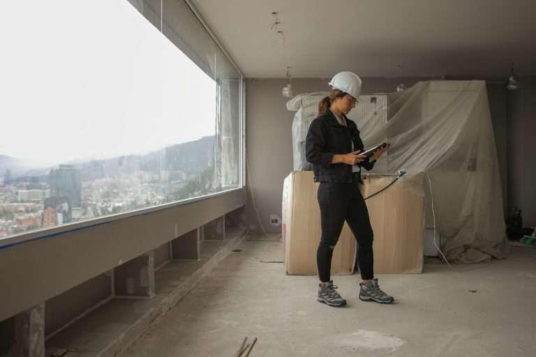 Qualitätskontrolle und Planung – Baureinigung Objektleiterin mit weißem Bauhelm und Tablet kontrolliert die Reinigungsarbeiten in einem Rohbau mit großen Fenstern.