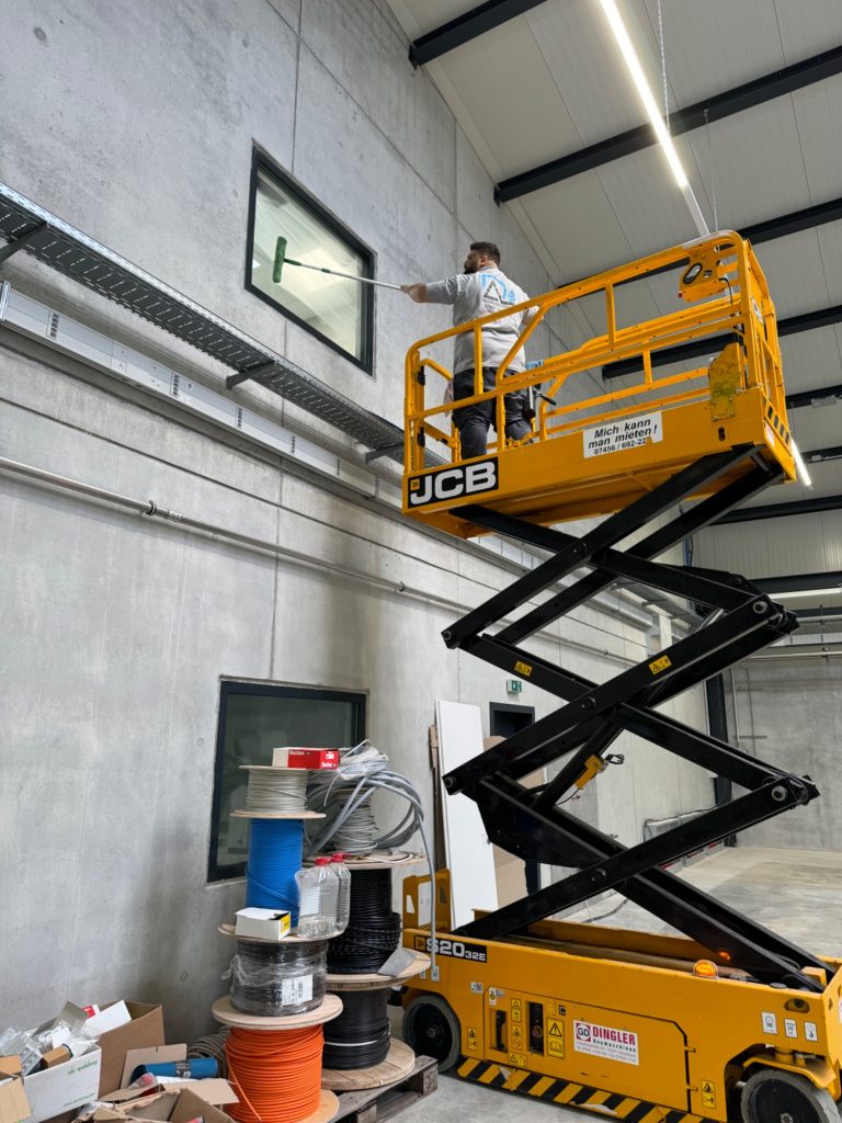 Fensterreinigung mit Hebebühne – Bauendreinigung Mitarbeiter bei der Fensterreinigung auf einer gelben Scherenarbeitsbühne in einer hohen Industriehalle während der Bauendreinigung.
