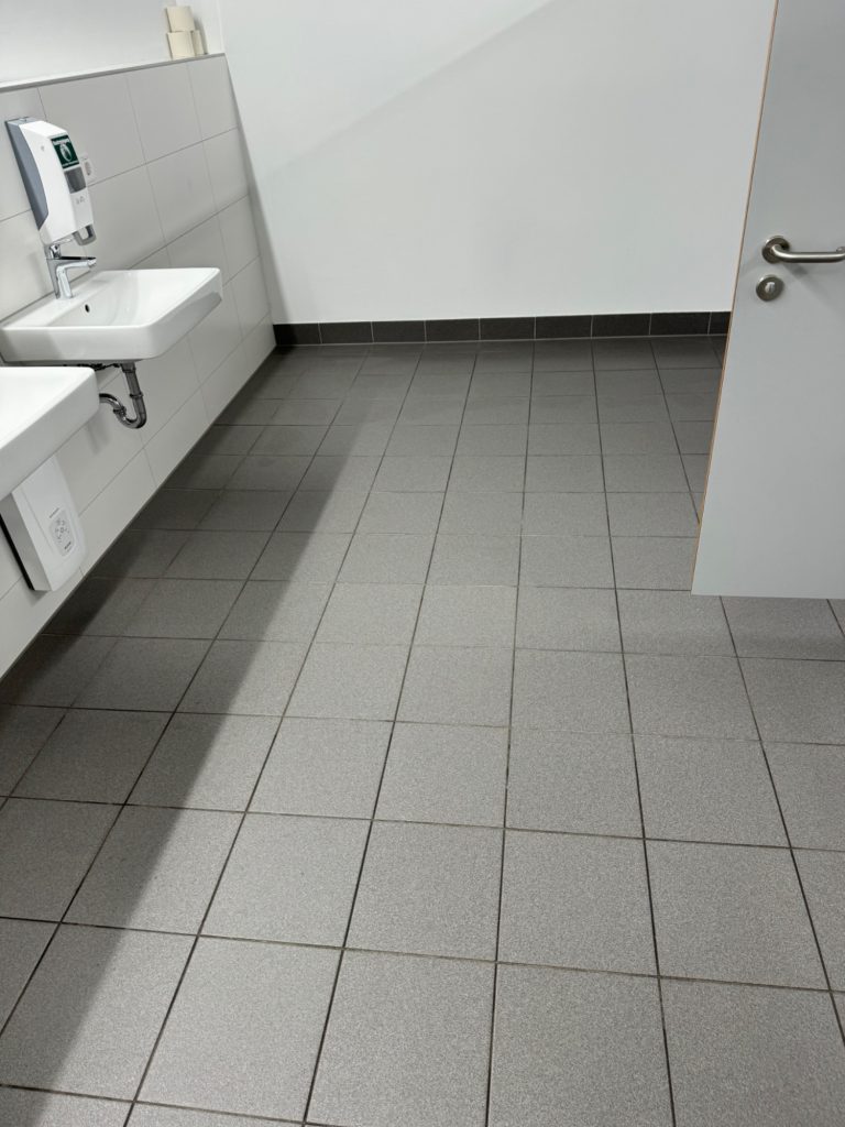 Ergebnis nach der Fliesen-Grundreinigung Sauberer, hellgrauer Fliesenboden nach der professionellen Grundreinigung in einem Sanitärraum.