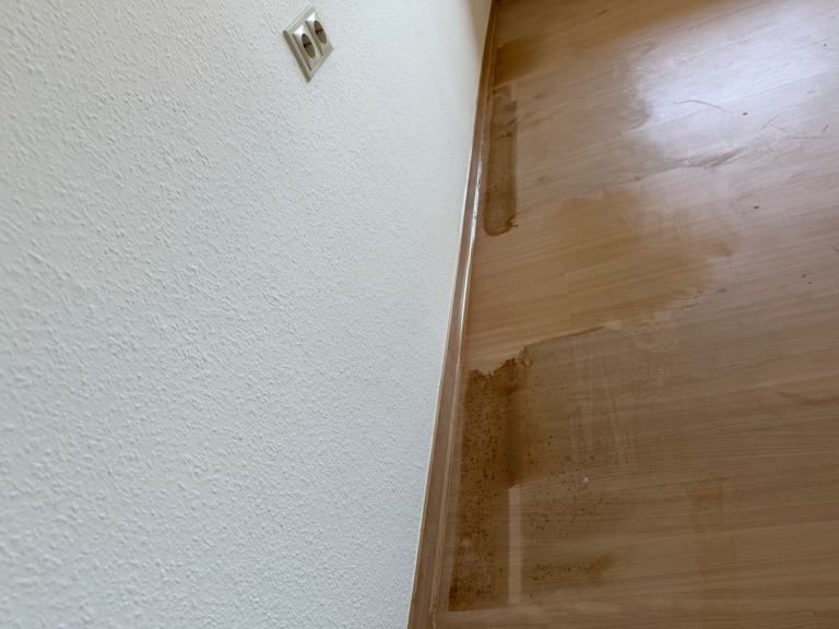 Vorher: Jahrelange Fettablagerungen auf Parkett Vorher-Ansicht eines Parkettbodens an einer Wandleiste mit extremen, dunkelbraunen Fettablagerungen aus einer ehemaligen Küche.